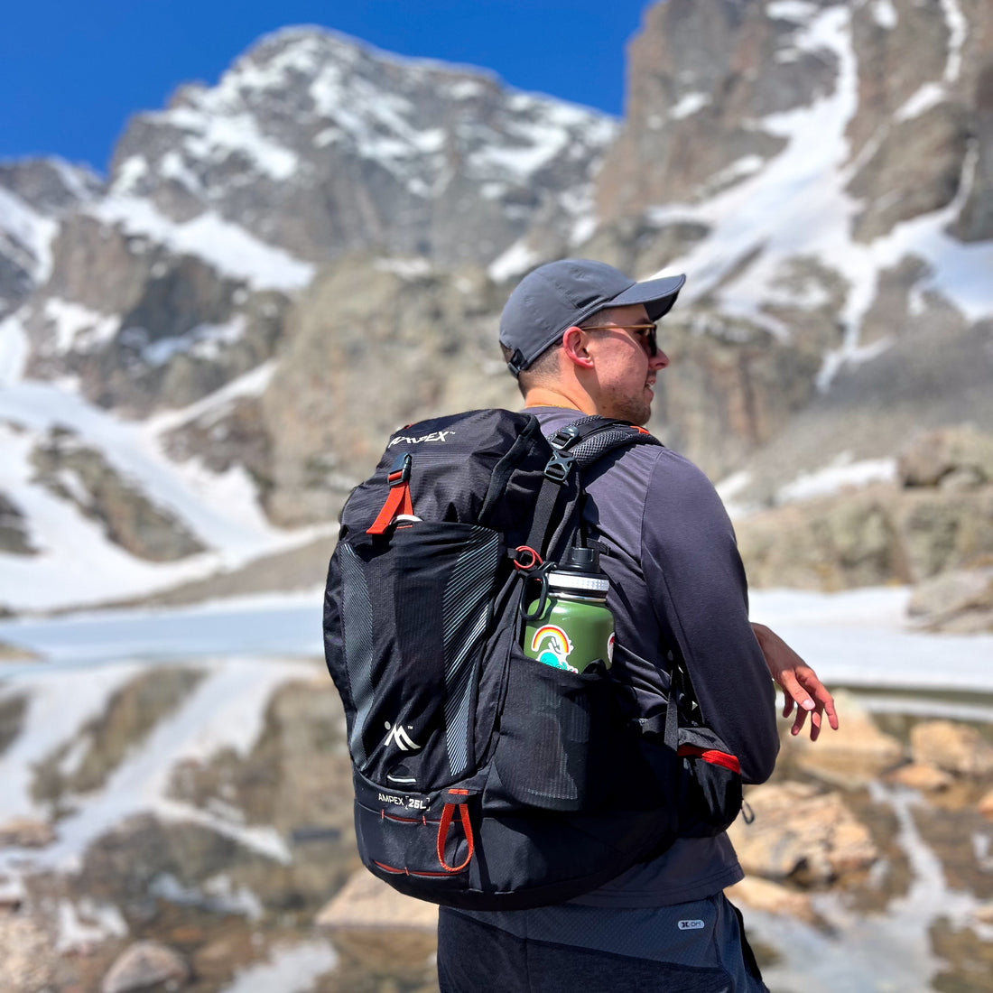 Person wearing the Ampex daypack while hiking near snowy mountain terrain, carrying a water bottle in the side pocket.