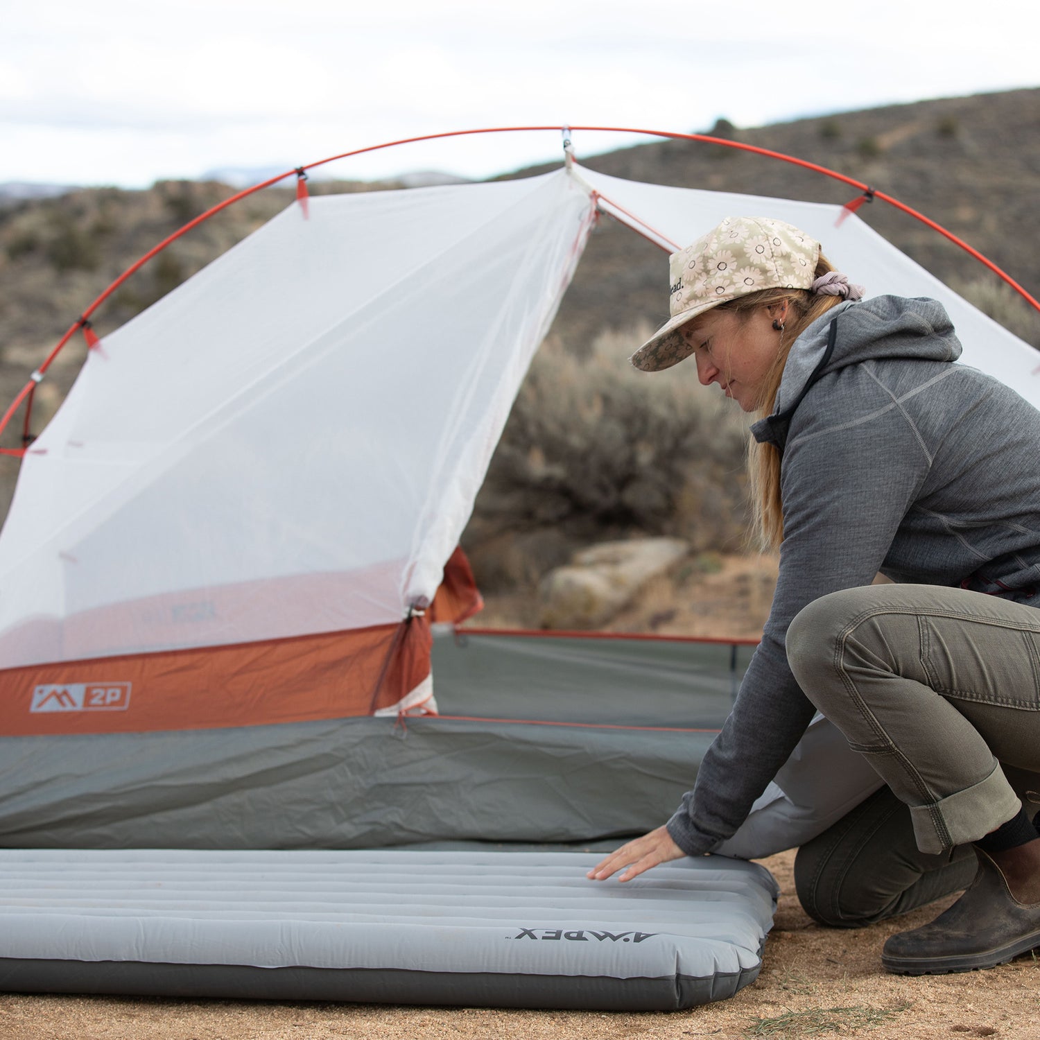 Person setting up a gray Ampex sleeping pad outside a tent in a campsite.