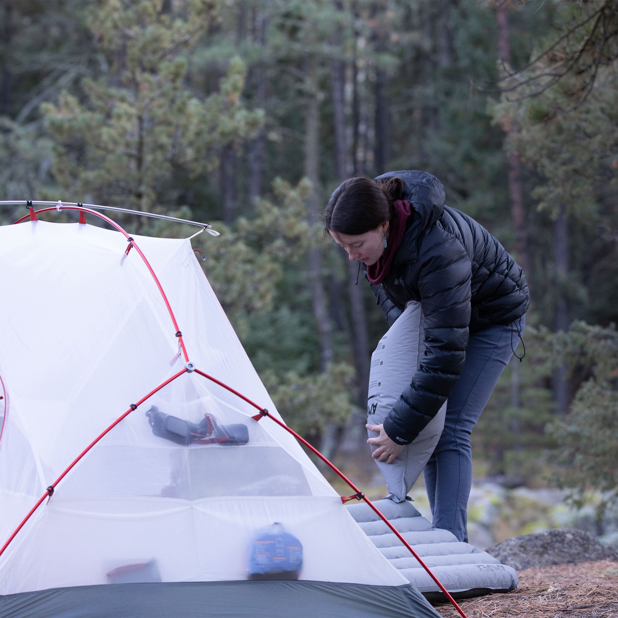 Person setting up a gray Ampex sleeping pad outside a tent in a forested campsite.