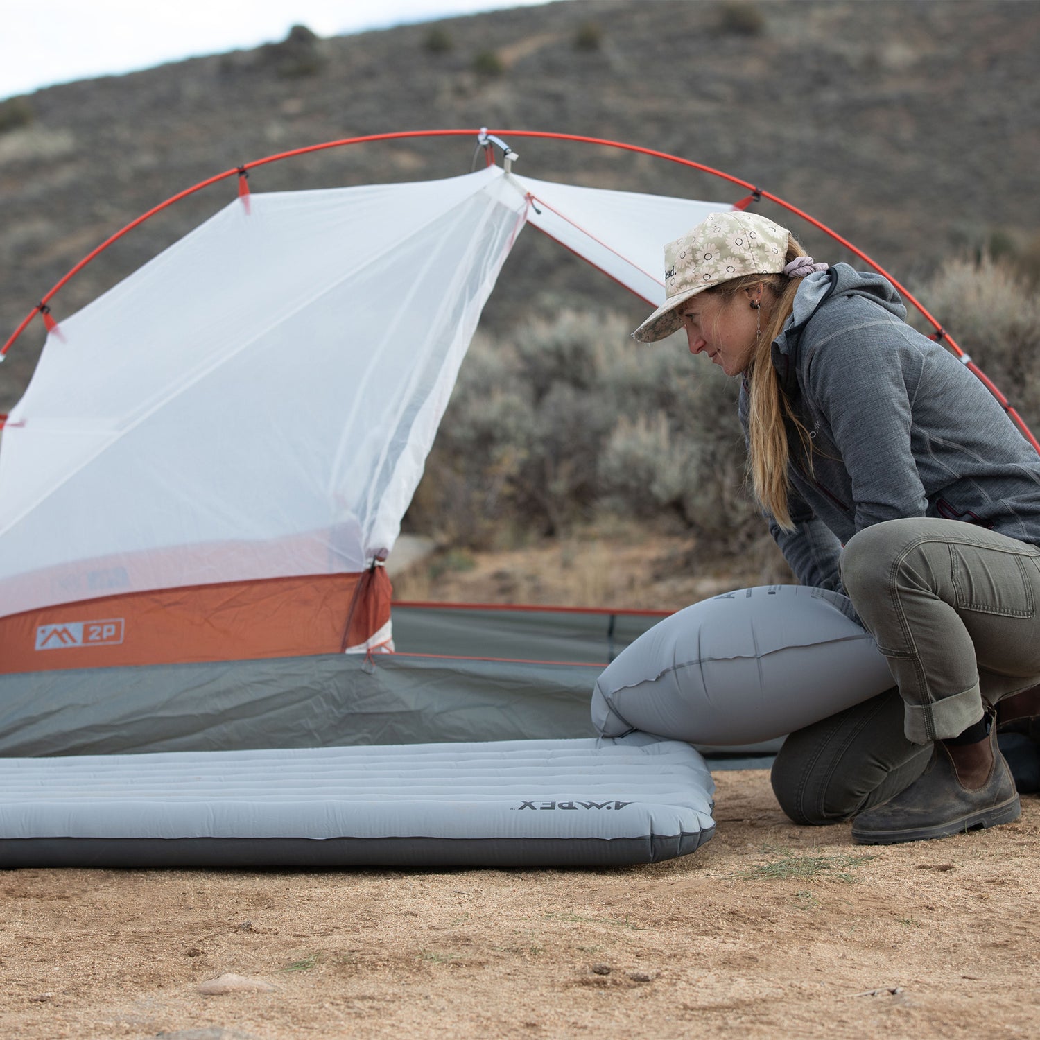 Person setting up a gray Ampex sleeping pad outside a tent in a campsite.