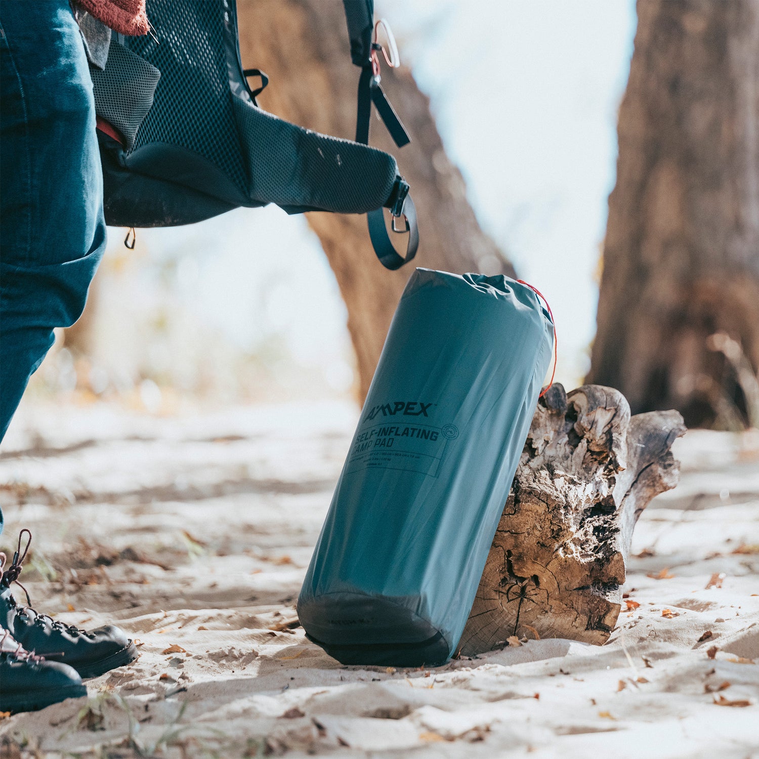 Person outdoors carrying the packed Ampex sleeping pad in its gray stuff sack with black straps.
