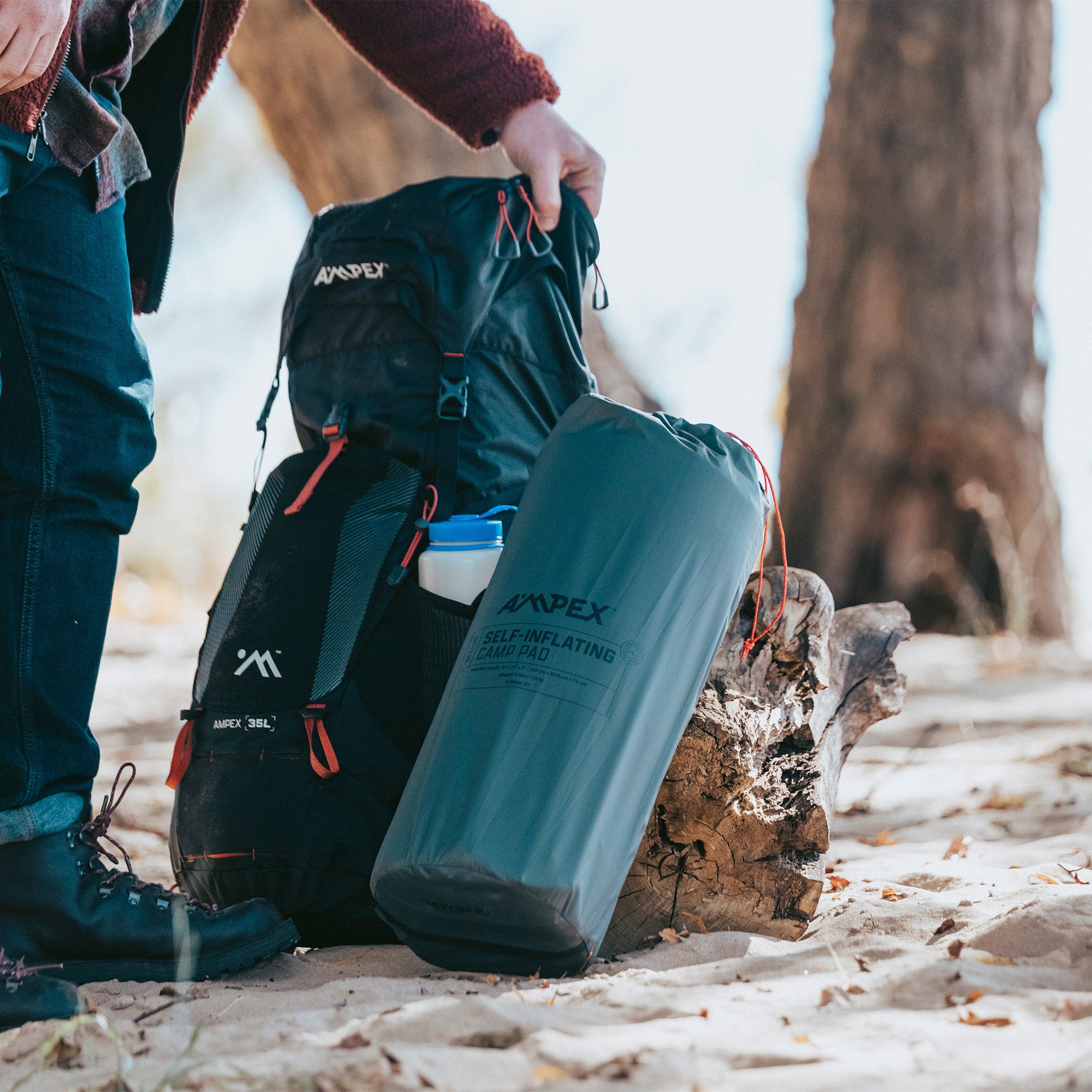 Person outdoors carrying the packed Ampex sleeping pad in its gray stuff sack with black straps.