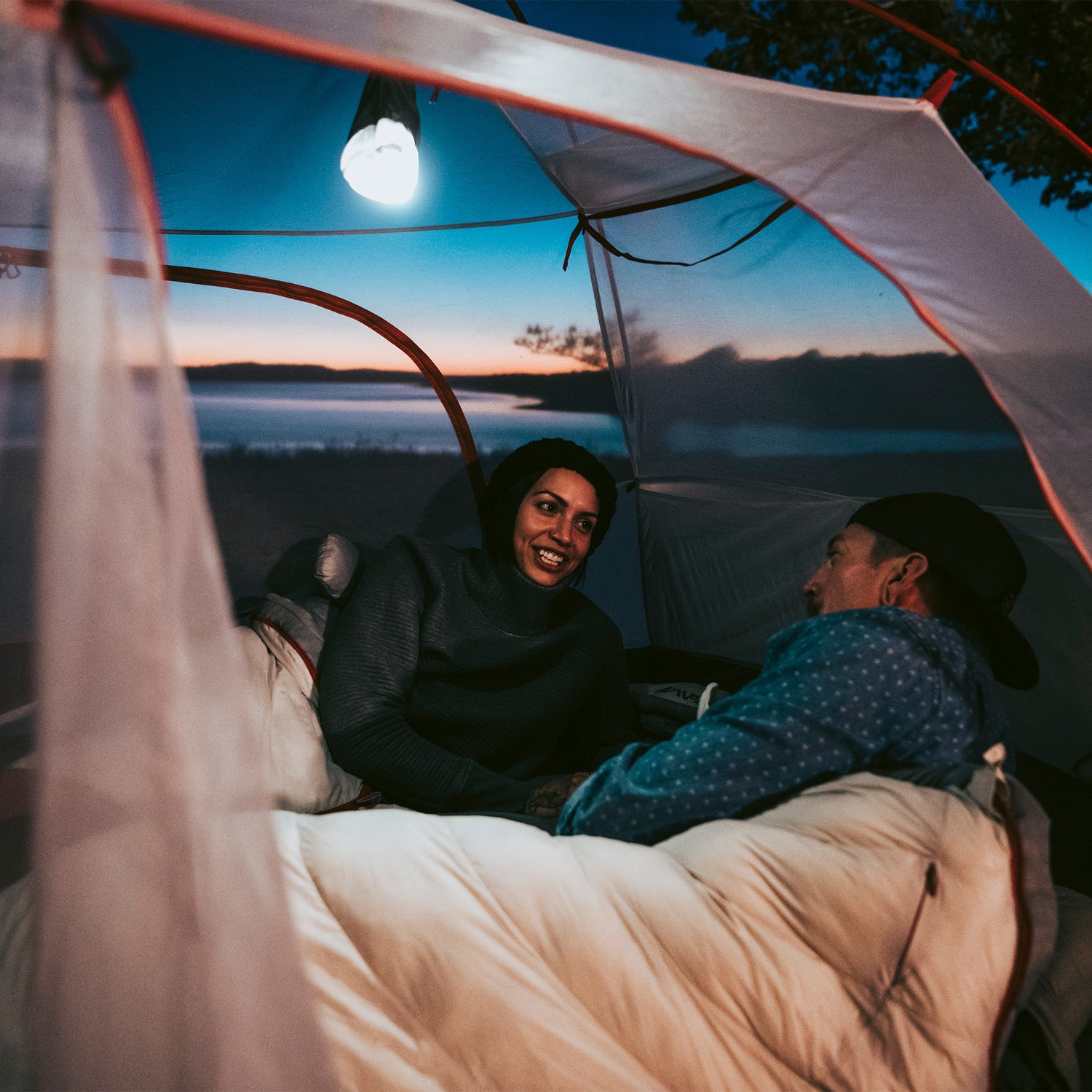 Two campers inside the tent at night, sitting in sleeping bags under the mesh canopy illuminated by a lantern.