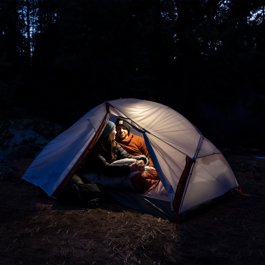 Two campers inside the tent at night, sitting in sleeping bags under the mesh canopy illuminated by a lantern.